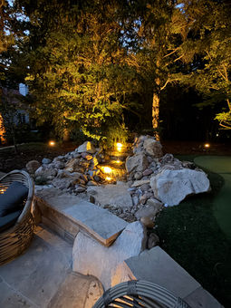 Illuminated rock waterfall feature in a backyard at night.