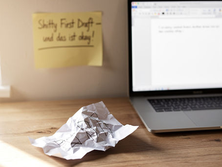 Ein zerknülltes Blatt Papier liegt auf einem Schreibtisch, daneben ein Laptop mit einem halbleeren Word-Dokument. Im Hintergrund ein Zettel mit der Aufschrift ‚Shitty First Draft – und das ist okay!‘. Das Bild symbolisiert den Befreiungsschlag vom Perfektionismus: Es geht nicht um Fehlerfreiheit, sondern um den Mut, einfach anzufangen.