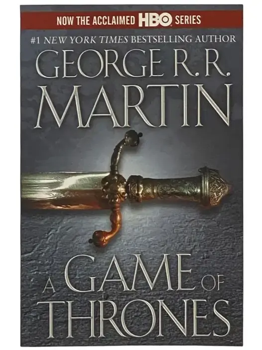 Book cover of A Game of Thrones by George R.R. Martin, featuring an intricate, detailed image of a silver sword. The sword is displayed prominently against a dark, medieval background, with ornate designs on the hilt and blade. The title A Game of Thrones is presented in bold, dramatic lettering at the top, with the series name A Song of Ice and Fire beneath it. The cover reflects themes of power, war, and the struggle for the Iron Throne, central to the epic fantasy series, with the silver sword symbolizing honor, conflict, and the deadly battles that lie ahead