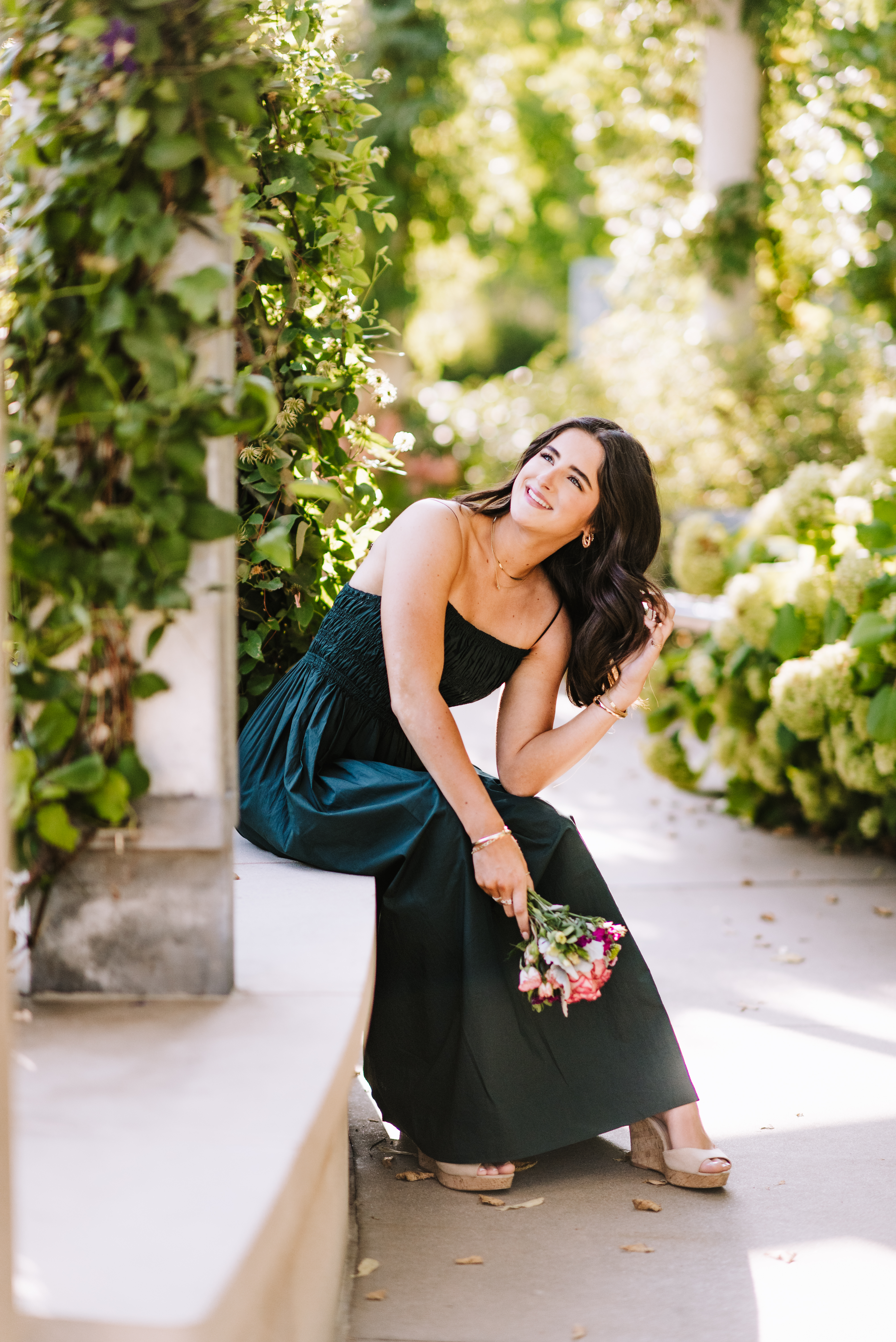 High school senior in a dark green dress holding flowers in a garden walkway