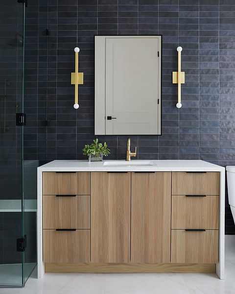 Contemporary bathroom with a fluted white oak vanity, waterfall quartz countertop, black tile accent wall, brass plumbing fixtures, and modern linear sconces flanking a framed medicine cabinet.