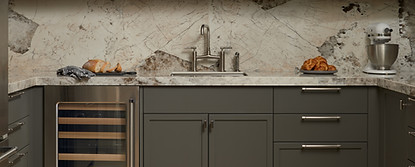 Modern transitional bar or prep kitchen with dark charcoal shaker cabinetry, open shelving, vertical shiplap wall detail, and a dramatic marble backsplash. Features include a built-in wine fridge, brushed metal hardware, light wood flooring, and styled decor with glassware, baskets, and ceramics.