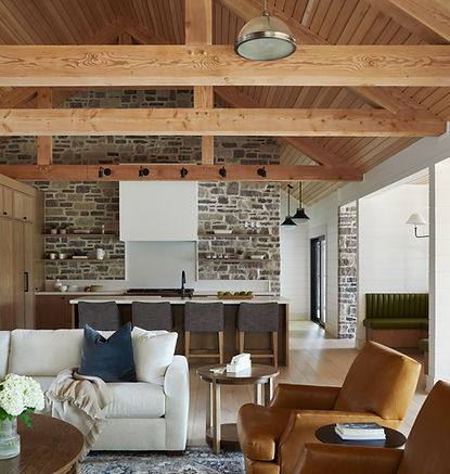 Rustic contemporary kitchen with warm wood cabinetry, exposed timber beams, and full-height stone walls. The space features a large waterfall island with seating for four, open wood shelving, a white range hood, black lighting accents, and vaulted ceilings with natural textures throughout.