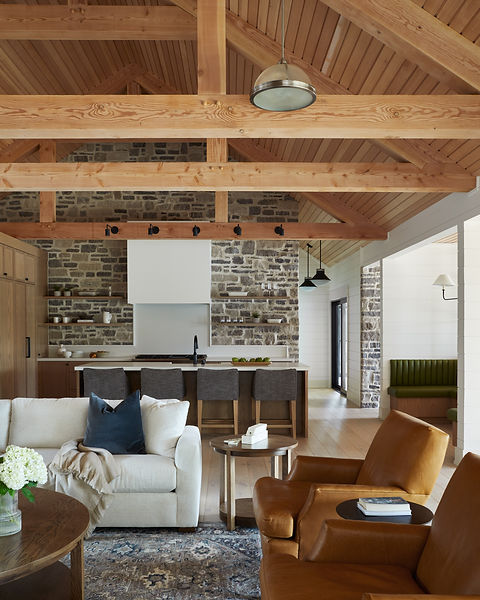 Rustic contemporary kitchen with warm wood cabinetry, exposed timber beams, and full-height stone walls. The space features a large waterfall island with seating for four, open wood shelving, a white range hood, black lighting accents, and vaulted ceilings with natural textures throughout.