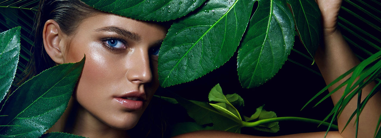 Beautiful model with professional soft glam makeup peering out from behind green foliage