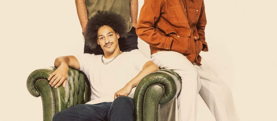 Three men pose against a beige background. One sits in a white shirt, another stands in a green shirt, and a third wears an orange jacket. Smiling mood.