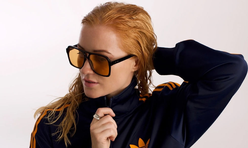 Woman with wet hair in a navy and orange track jacket adjusts oversized sunglasses, exuding a confident and stylish mood against a plain backdrop.