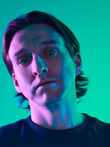 Man with neutral expression in a blue and green-lit setting. He wears a dark shirt, hair slightly long, and stands against a smooth gradient background.