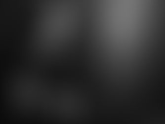 Man seated in dim room with dark walls and exposed concrete. Neutral expression. Black and white tone. Electrical cord visible.