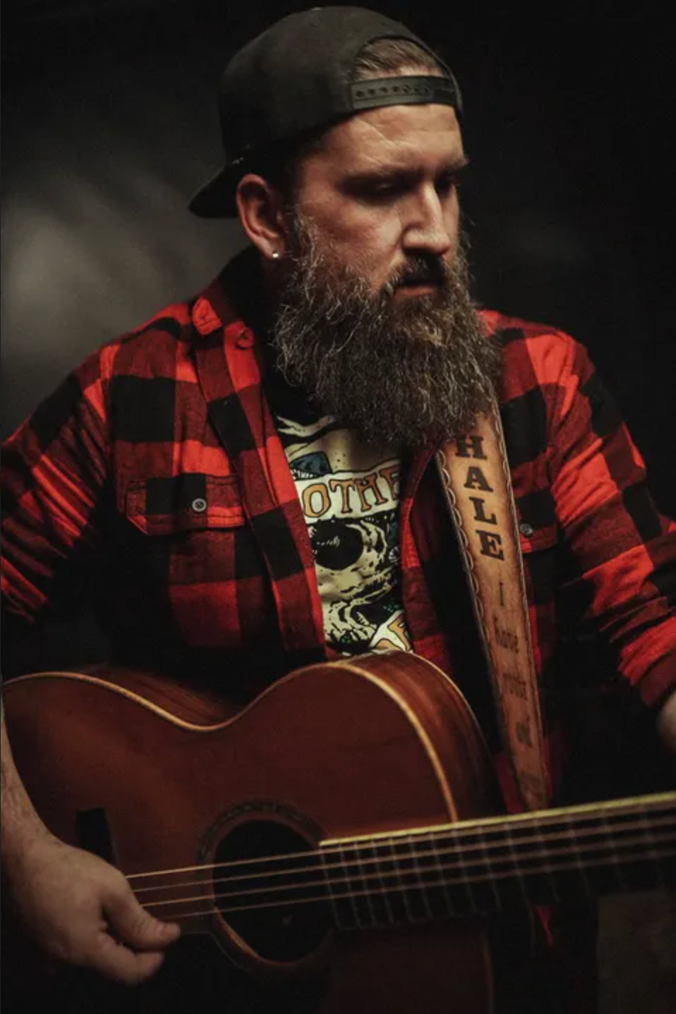 Bearded man in a backward cap and red plaid shirt plays an acoustic guitar against a dark background, with a HALE strap.