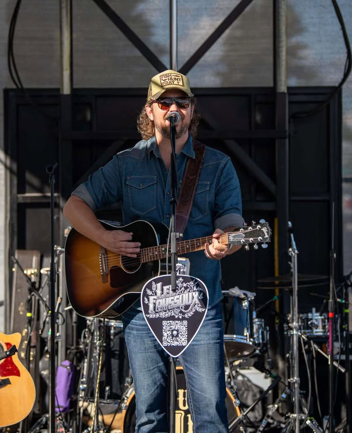 Country musician sings and plays acoustic guitar on an outdoor stage, wearing a cap and sunglasses, with a drum kit behind.