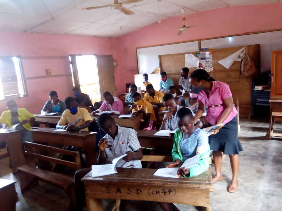 Victoria Aggrey is in her classroom with students sitting at small desks, reading from the books, some with facemasks on. Victoria is in a pink blouse and grey skirt, leaning over to help one of her students. The classroom has pink walls, a couple of fans, a white board, and cement floors.