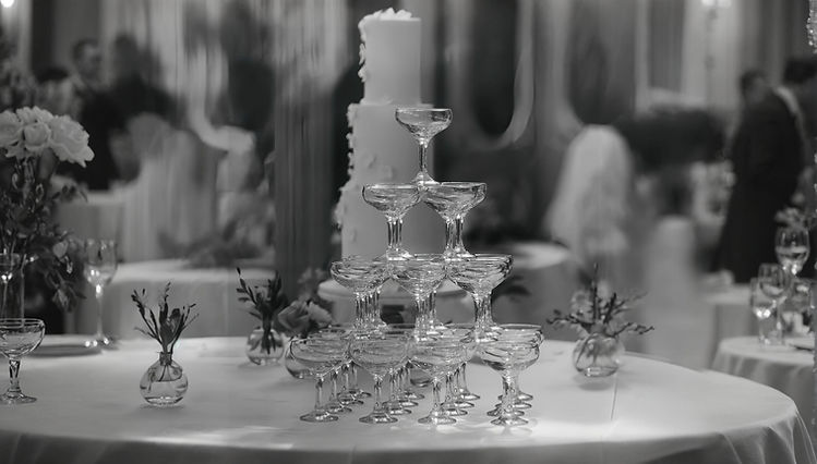 A Black and White image showing aThree-Tiered cake with a sugar blossom design standing behind a Champagne Tower
