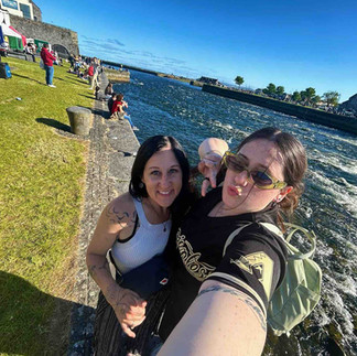 Mara and Lily looking up at the camera in front of a large river; Mara smiling and Lily pursing her lips in a kiss