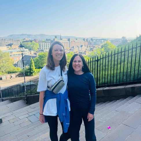 Two white women with long dark hair smiling at the camera, standing on the steps in front of Edinburgh city