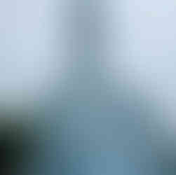 A tall, light-blue church with ornate white trim and a clock tower topped by a cross, photographed from below against a cloudy sky, with a few people and parked cars at street level.