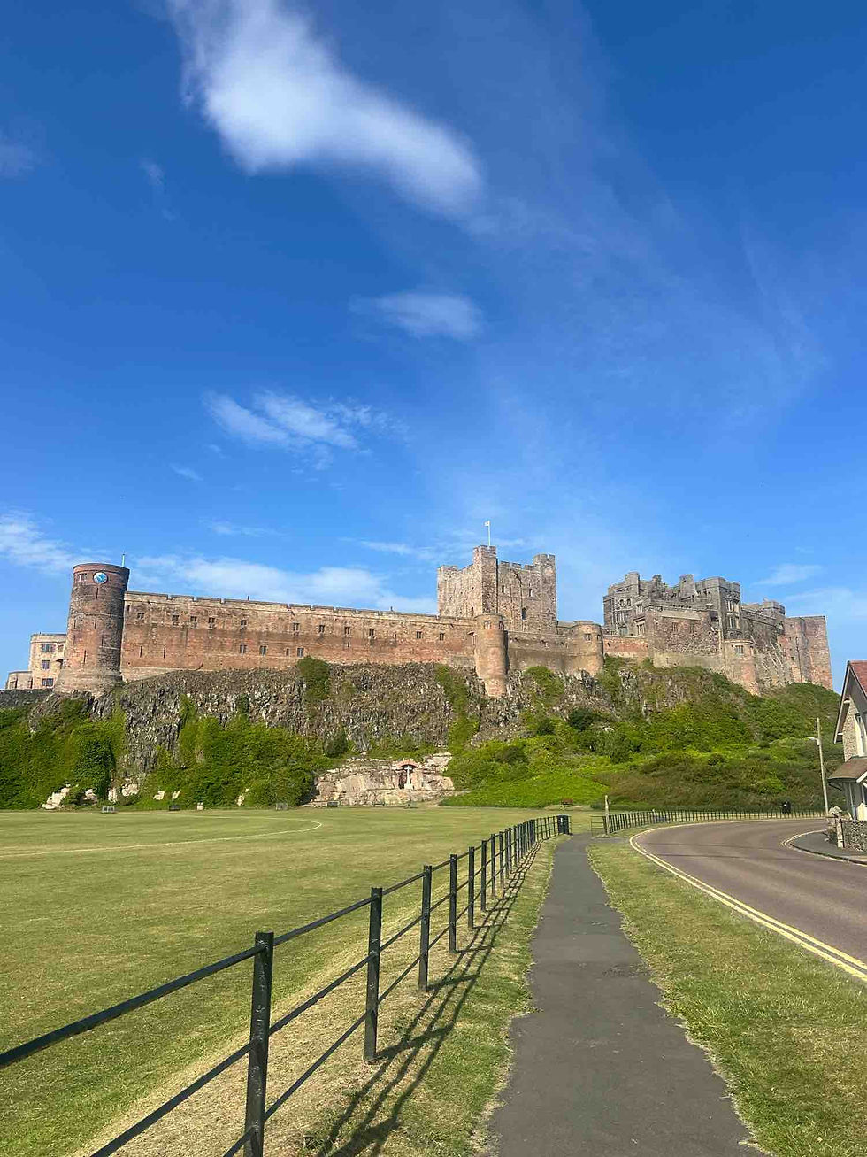 A narrow sidewalk running between the road and an iron fence next to an open green field, views of a large medieval stone castle ahead