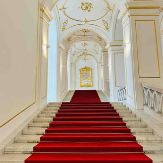 Inside a castle stairway that is cloaked in red velvet carpet, white arches walls and ceilings trimmed with gold.