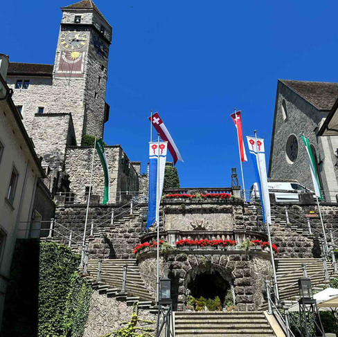 Stone steps that wrap around either side of a large stone arch adorned with bright red flowers and Swiss flags, leading up to a large medieval clock tower under a bright blue sky.