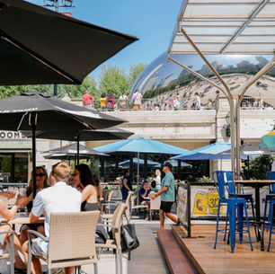 People dine under umbrellas at a sunny outdoor patio, with the reflective Cloud Gate sculpture and crowds in the background.