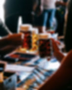 Hands clink beer mugs over a table with menus, in a lively setting. Warm lighting and amber hues create a cheerful atmosphere.