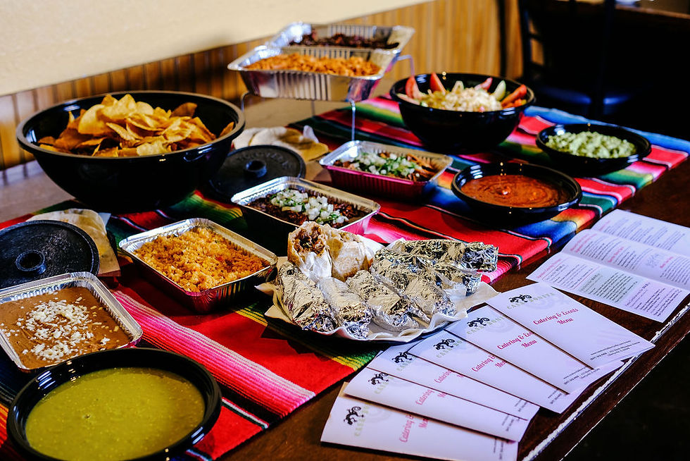 A table with various Mexican dishes like tacos and dips, bright striped tablecloth, and catering menus in a cozy setting.