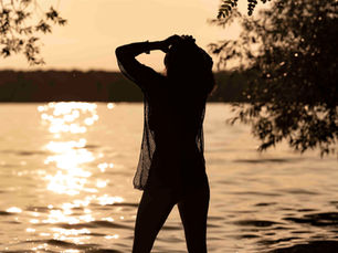 A woman in a sensual, relaxed pose gazes thoughtfully over a calm lake, surrounded by nature. Her posture radiates softness, presence, and feminine energy, capturing a moment of stillness and inner connection.