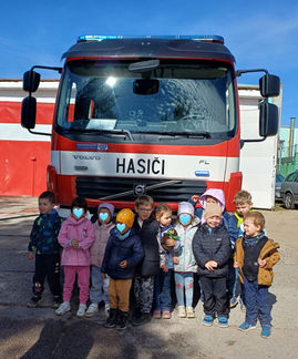 Děti ze soukromé školky stojí společně před hasičským autem během exkurze u dobrovolných hasičů a s nadšením poznávají práci záchranářů.