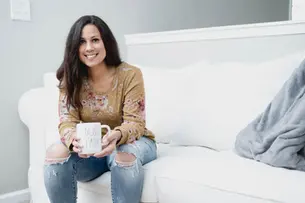 Crystal Waddell, in a floral sweater, sits on a white couch, smiling and holding a mug labeled "Boss Lady." Cozy setting with a gray blanket.