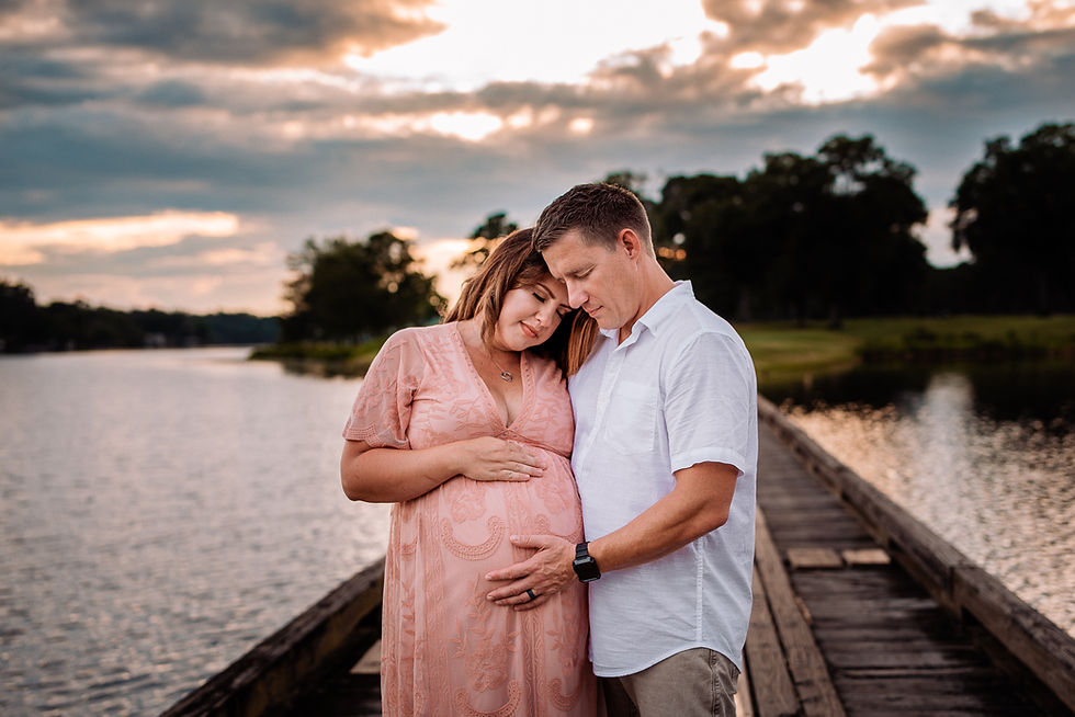 Couple embraces on a wooden dock by a lake at sunset. The woman in a peach dress cradles her belly, while the man in white holds her hand for maternity photos.