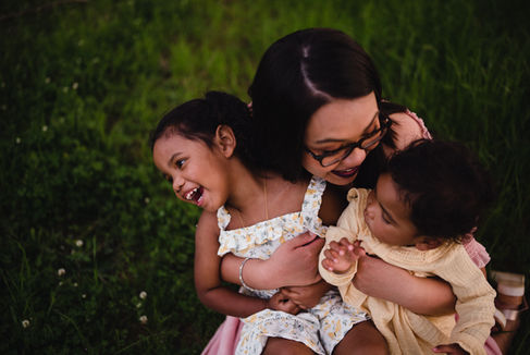 Mom tickles daughters while they are sitting in her lap in tall grass