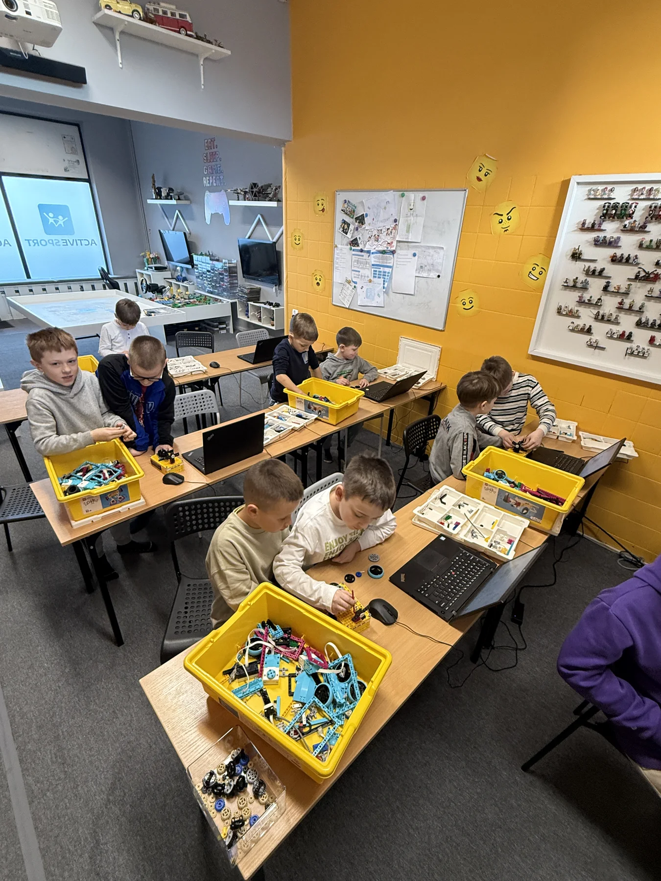 Dzieci budują konstrukcje LEGO przy stołach z laptopami i zestawami klocków