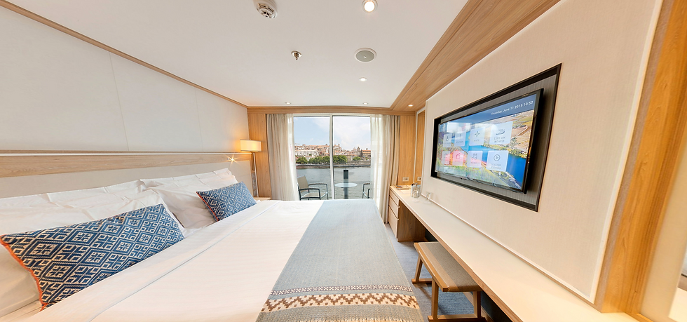 Hotel room with a neatly made bed, blue patterned pillows, and a wall-mounted TV showing a menu. Large window with a river view. Calm ambiance.