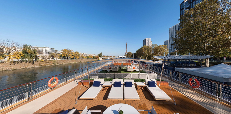 Sunlit riverboat deck with empty lounge chairs, Eiffel Tower visible in distance. Trees and cityscape line the river. Clear blue sky.