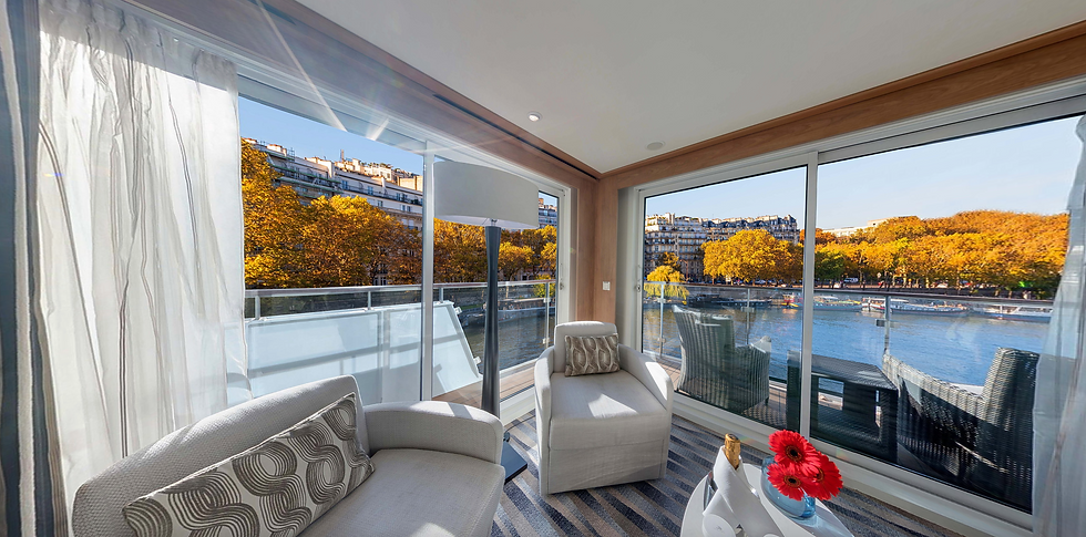 Modern room with beige furniture and a vase of red flowers. Large windows offer a view of a river and trees with autumn foliage outside.