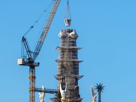 Crane hoisting a structure atop a scaffolding-covered tower against a clear blue sky. Decorative elements with star motifs are visible.