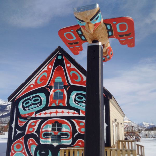 Colorful totem art on a house facade with a red, black, and blue pattern. A carved bird sits atop a post. Snowy mountain backdrop