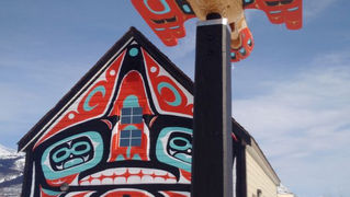 House with vibrant Indigenous art features red, black, blue patterns. A totem-like structure stands in front. Snowy background, clear sky.