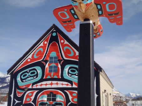 House with vibrant Indigenous art features red, black, blue patterns. A totem-like structure stands in front. Snowy background, clear sky.