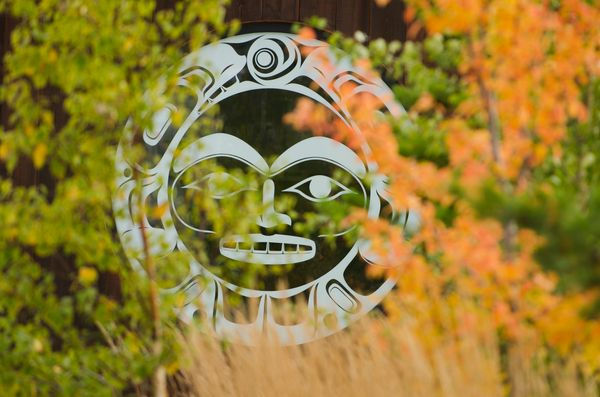 A white sun mask design is visible through vibrant autumn foliage. The setting is outdoors, creating a mysterious and serene mood.