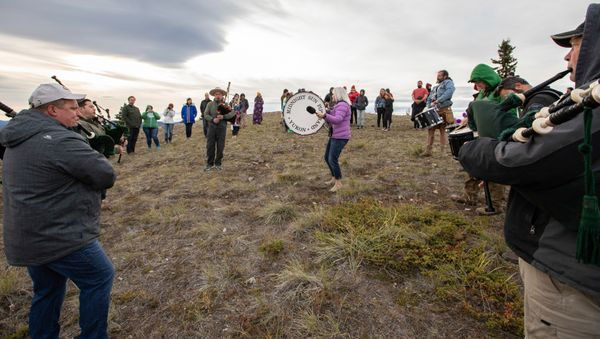 A group plays bagpipes and drums on a grassy hillside under a cloudy sky. Participants wear jackets in various colors, creating a lively scene.