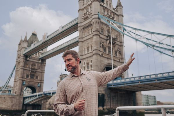 Man with a headset gestures towards Tower Bridge in London. He's wearing a checked shirt. The sky is cloudy, creating a neutral mood.
