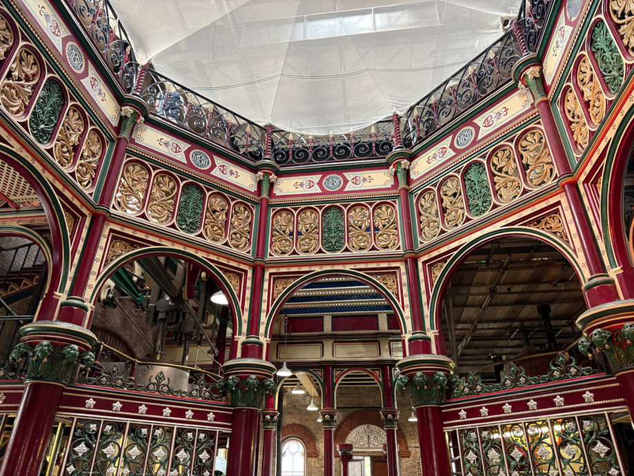 Ornate red, green, and gold Victorian-style market hall interior with arches, iron railings, and MBM medallions under a canopy.