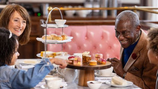 Family enjoying tea with cakes in a cozy café. There is a pink cushioned booth and a tiered tray with pastries on the table. Smiling faces.