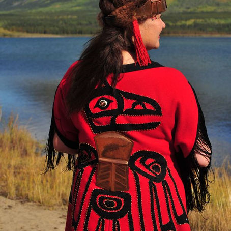 A person in a red ceremonial robe with black tribal designs stands by a lake, with mountains in the background. Sunny day.