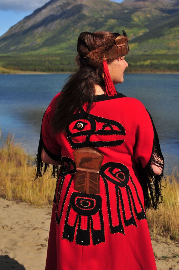 A person in a red ceremonial robe with black tribal designs stands by a lake, with mountains in the background. Sunny day.