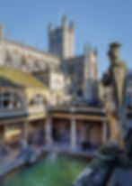 Historic architecture with detailed stonework surrounds a steaming bath. Statues overlook tourists exploring the site under a clear sky.