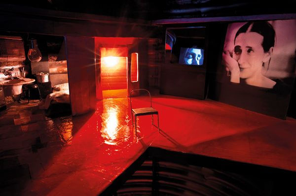 Dimly lit room with red light, featuring a chair and a tub. Walls show large portraits of a woman. Mysterious, contemplative mood.