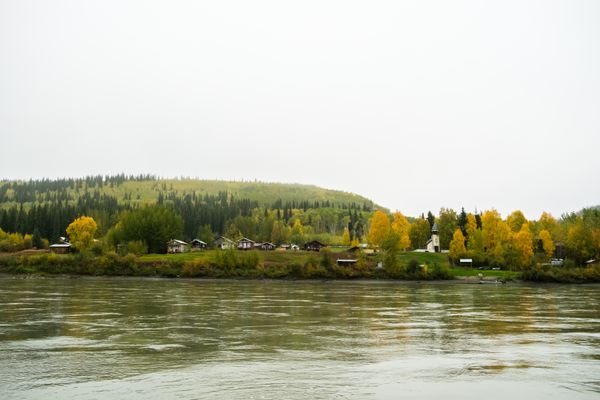 A tranquil riverside scene with a village of houses and a church among autumn trees under a cloudy sky. Hills and forest in the background.