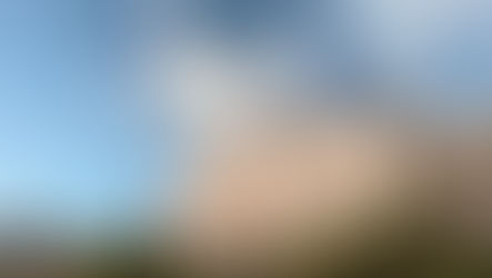 Sunlit stone building labeled "VISTA" under a blue sky with clouds. Surrounded by greenery, palm trees, and a distant historic structure.